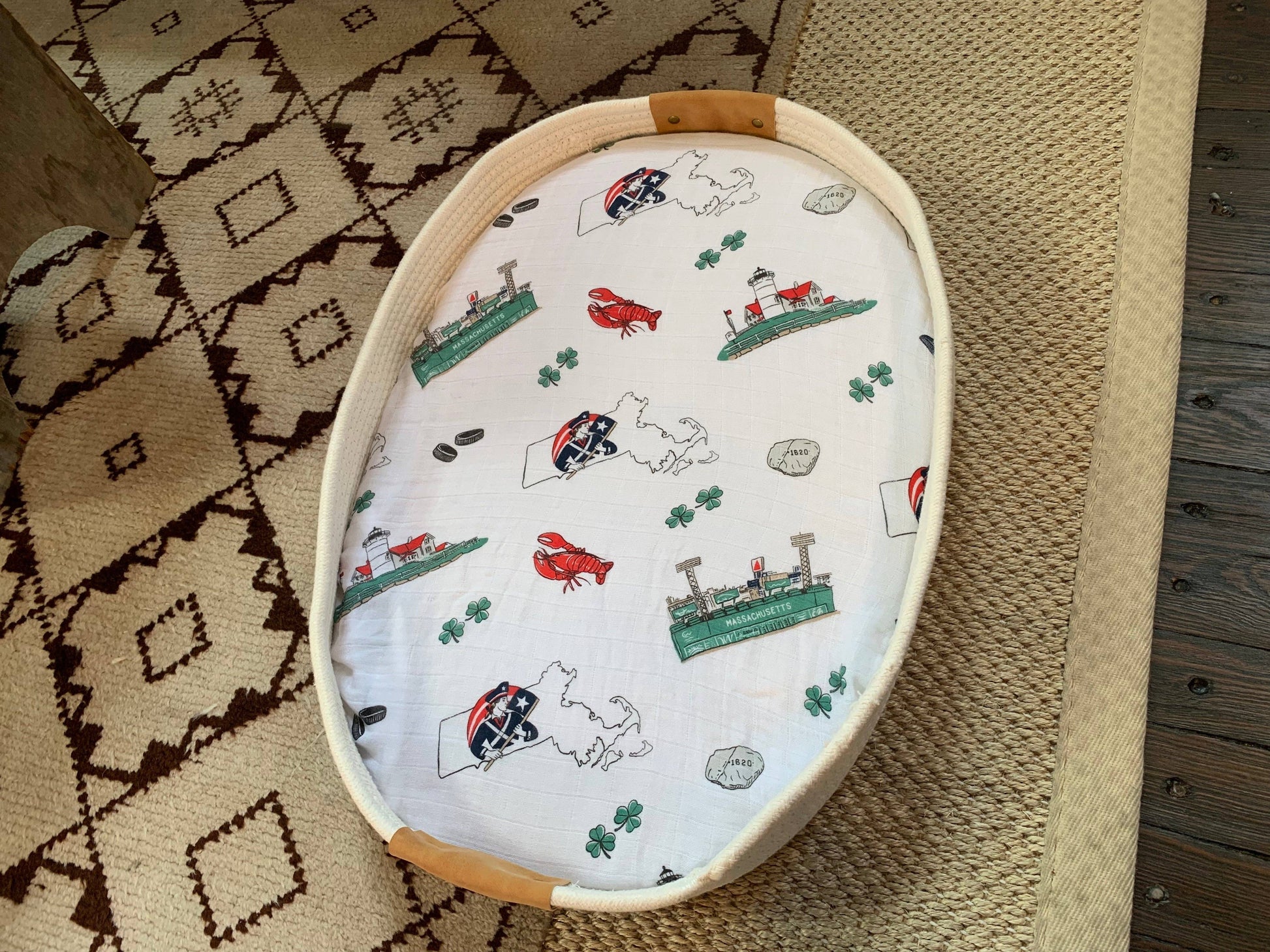 Mellow Monkey Massachusetts themed muslin baby swaddle blanket with state icons in a rope basket on a patterned rug.