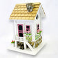 Mellow Monkey May Cottage birdhouse with white exterior, pink door and windows, stone accents, and floral details.