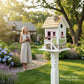 Mellow Monkey May Cottage birdhouse with white exterior, pink door and windows, flower boxes, stone accents, and wood shingled roof.