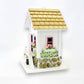 Mellow Monkey May Cottage birdhouse with white exterior, pink window frames, flower boxes, stone base, and wooden shingle roof.