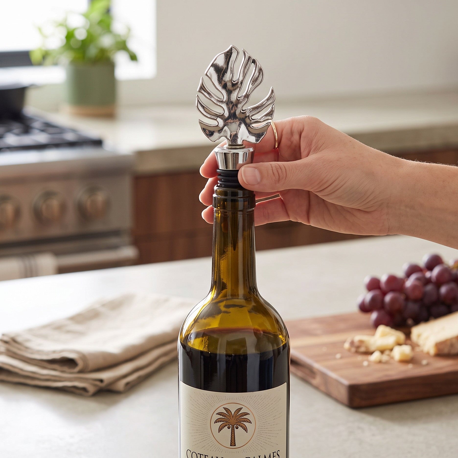 Mellow Monkey silver monstera leaf wine bottle stopper being placed into a wine bottle on a kitchen counter.