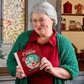 Mellow Monkey Mrs. Claus 12 Days of Cookies recipe book held by a woman dressed as Mrs. Claus in a festive kitchen.