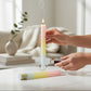Mellow Monkey multi-scented pink, yellow, and blue square taper candle being lit by hand on a white table.