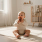 Baby holding a brown natural rubber Mellow Monkey pacifier while sitting on a rug in a bright nursery