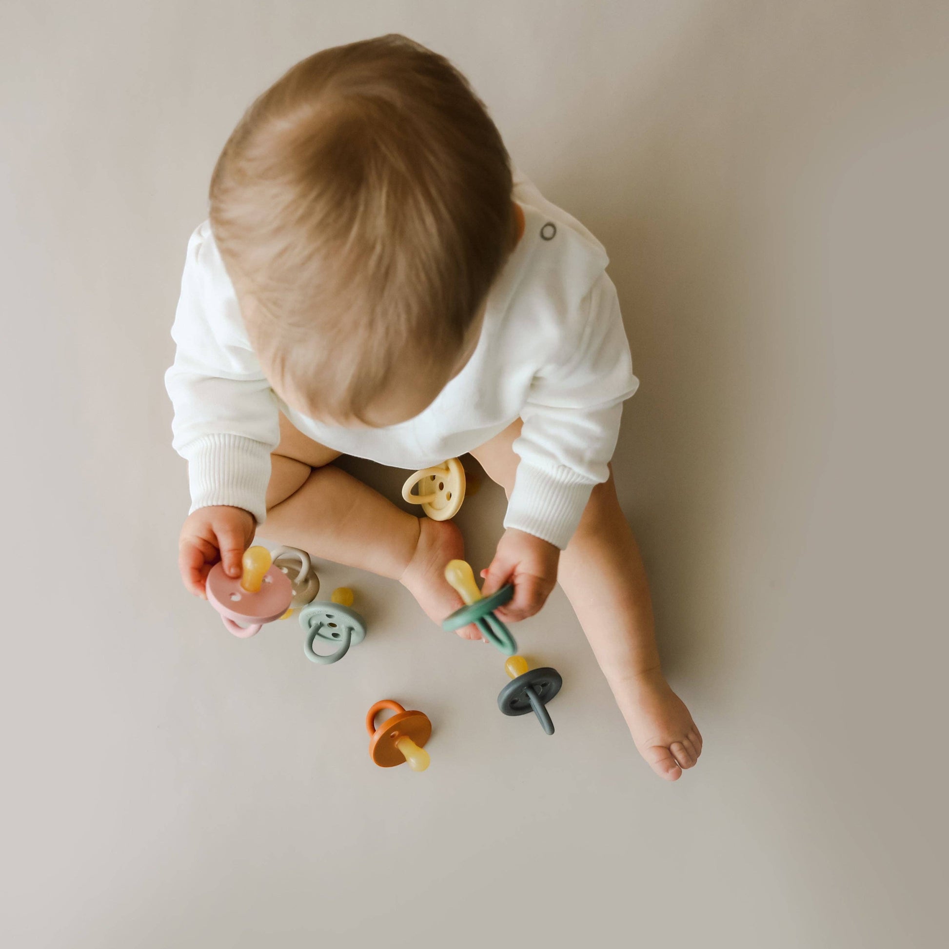 Mellow Monkey eco-friendly natural rubber pacifiers in assorted muted colours held by a baby in a white long-sleeve top