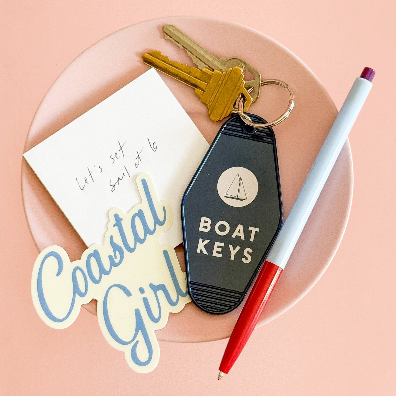 Mellow Monkey retro-style navy blue boat keys keychain with white sailboat icon on a pink plate with a pen and sticker.