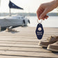 Mellow Monkey navy blue retro motel-style Boat Keys keychain held over a wooden dock with a sailboat in the background.