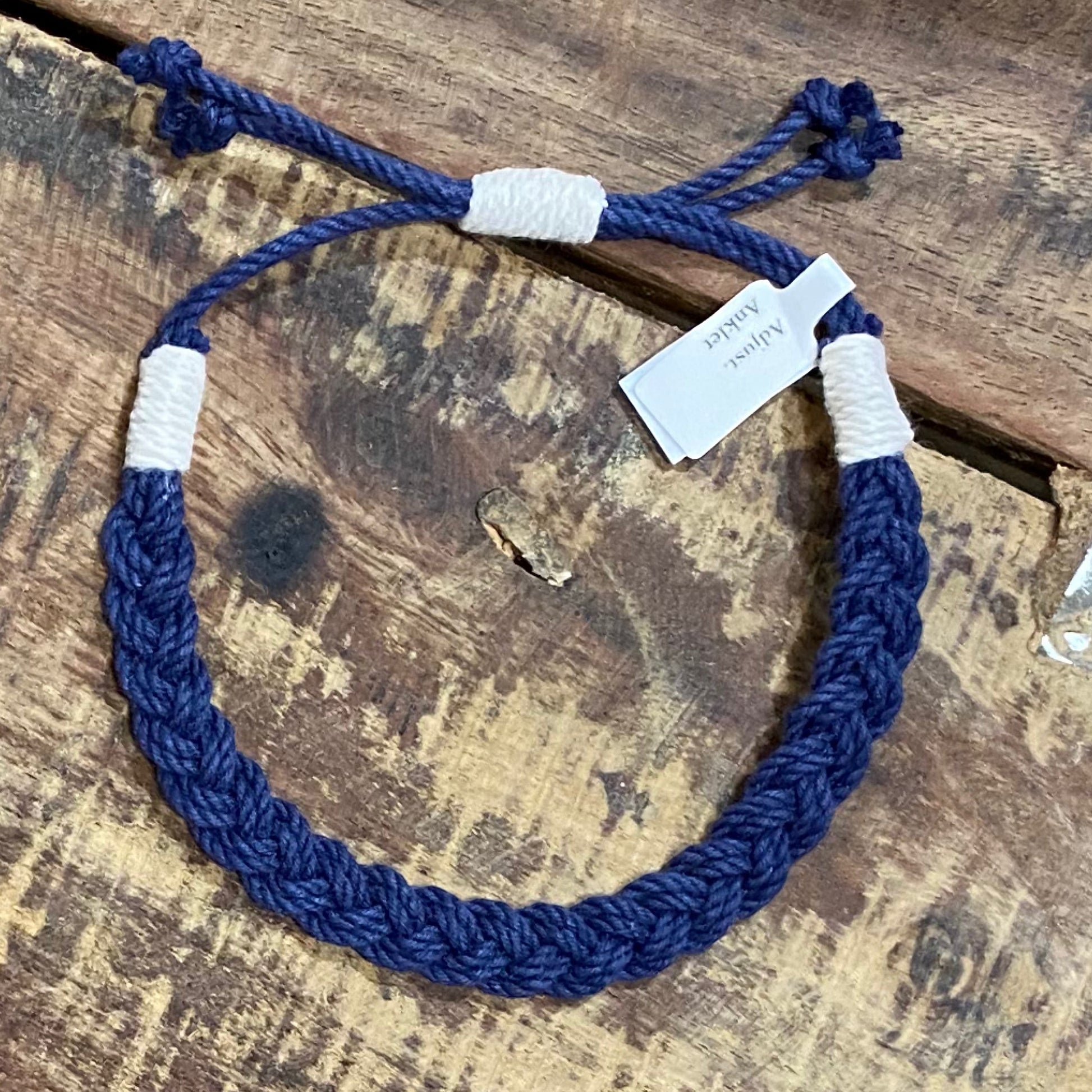 Mellow Monkey navy blue woven cotton nautical anklet with adjustable white cord knots on a rustic wooden surface