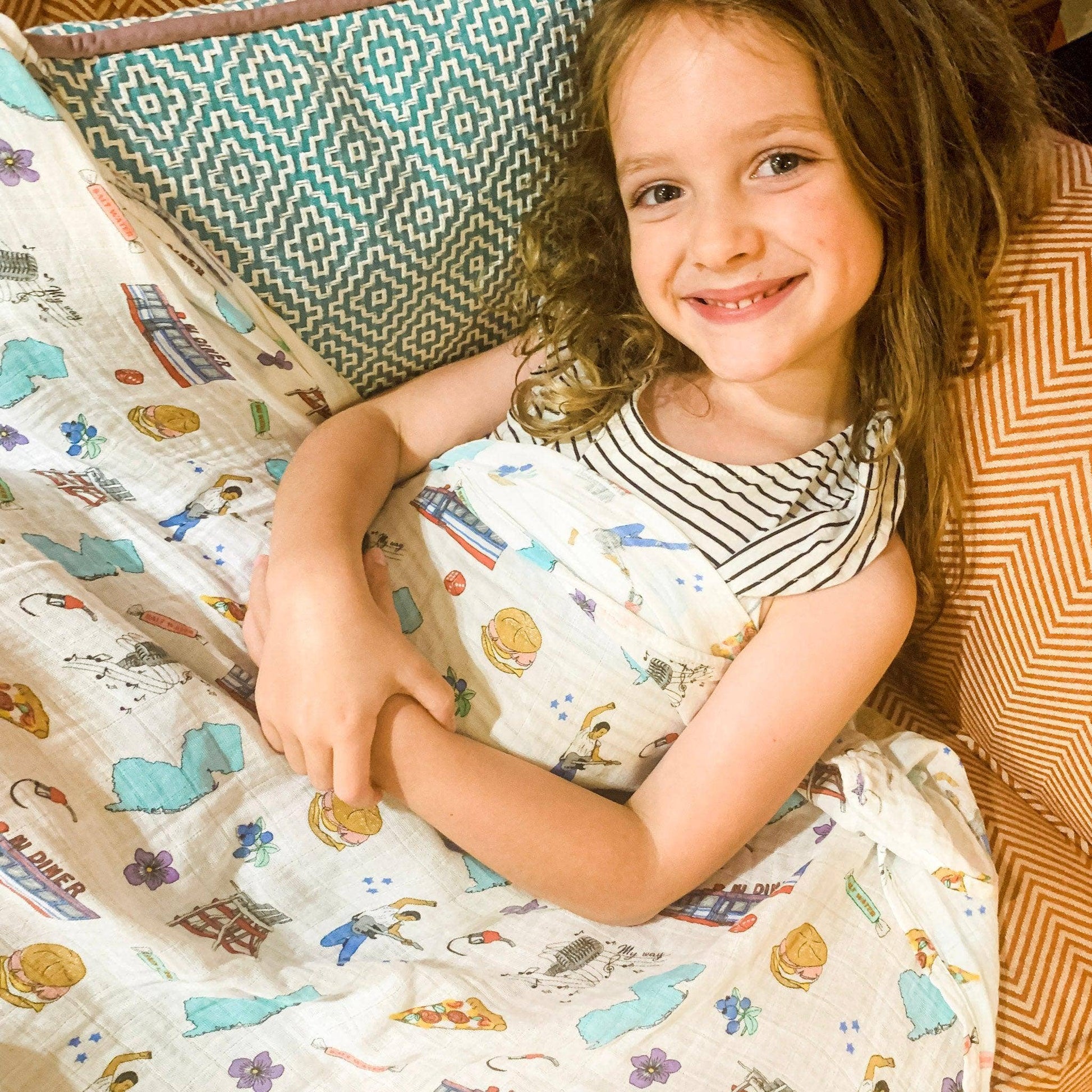 Mellow Monkey New Jersey themed muslin swaddle blanket featuring state icons, covering a smiling young girl.