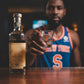 Mellow Monkey New York Knicks 1.7oz clear glass shot glass held by a man wearing a Knicks jersey in a home bar setting.