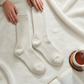 Mellow Monkey oatmeal ragg crew socks with a textured knit design, displayed on a soft white blanket with a book.