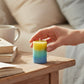 Mellow Monkey multi-scented pillar candle with yellow, green, and blue ombre layers on a wooden bedside table.