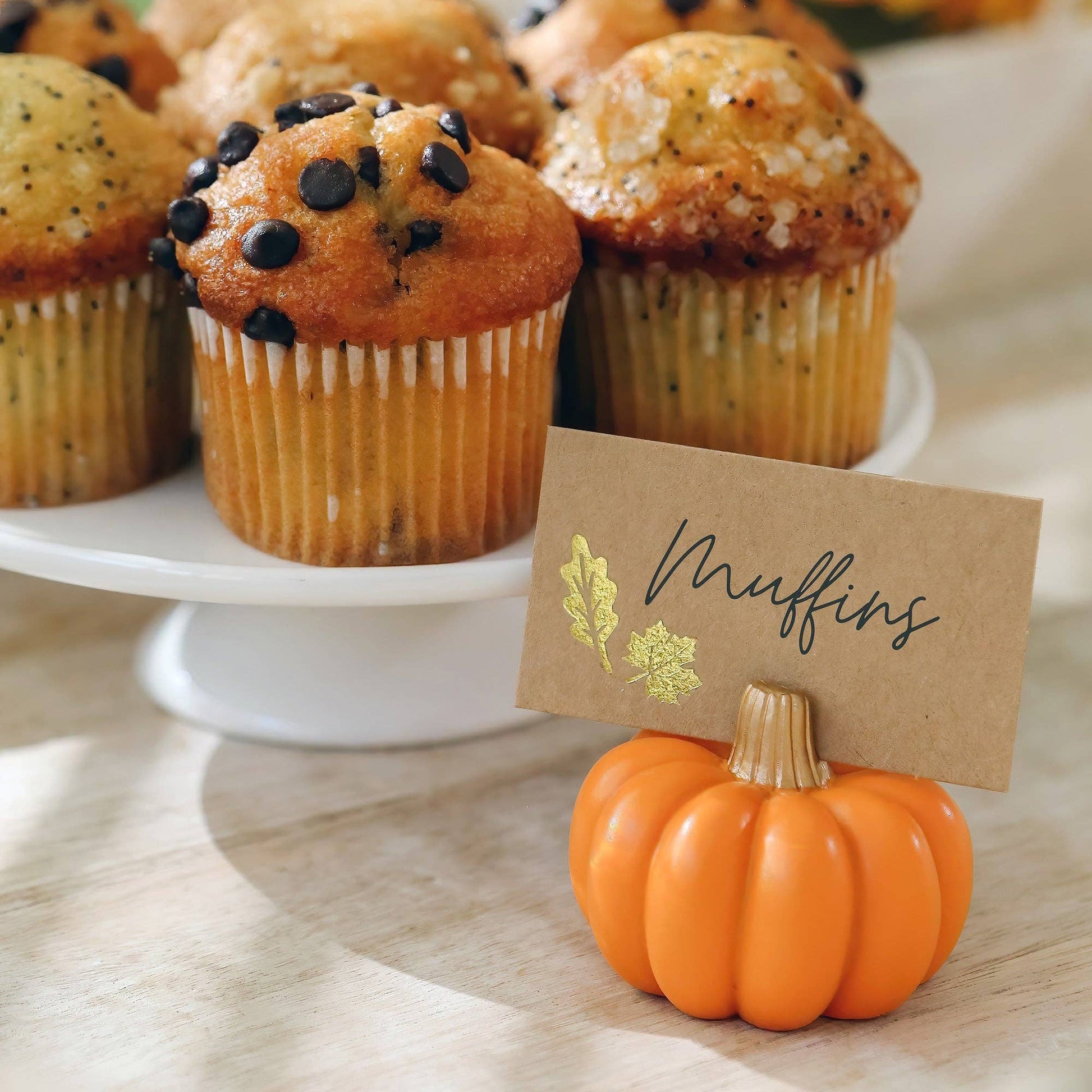 Mellow Monkey orange pumpkin place card holder with a gold-embellished card in front of a tray of muffins