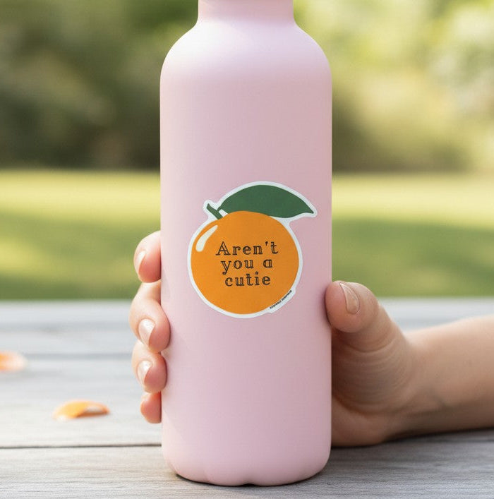Mellow Monkey Aren't You A Cutie vinyl orange sticker on a light pink water bottle held by a hand outdoors