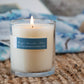 Mellow Monkey 9-ounce soy blend candle in a clear glass jar with a blue label, lit and glowing on a textured mat.