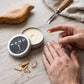 Mellow Monkey Wood Aromatics Palo Santo hand salve in a 2 oz tin with a person applying cream on a wooden table