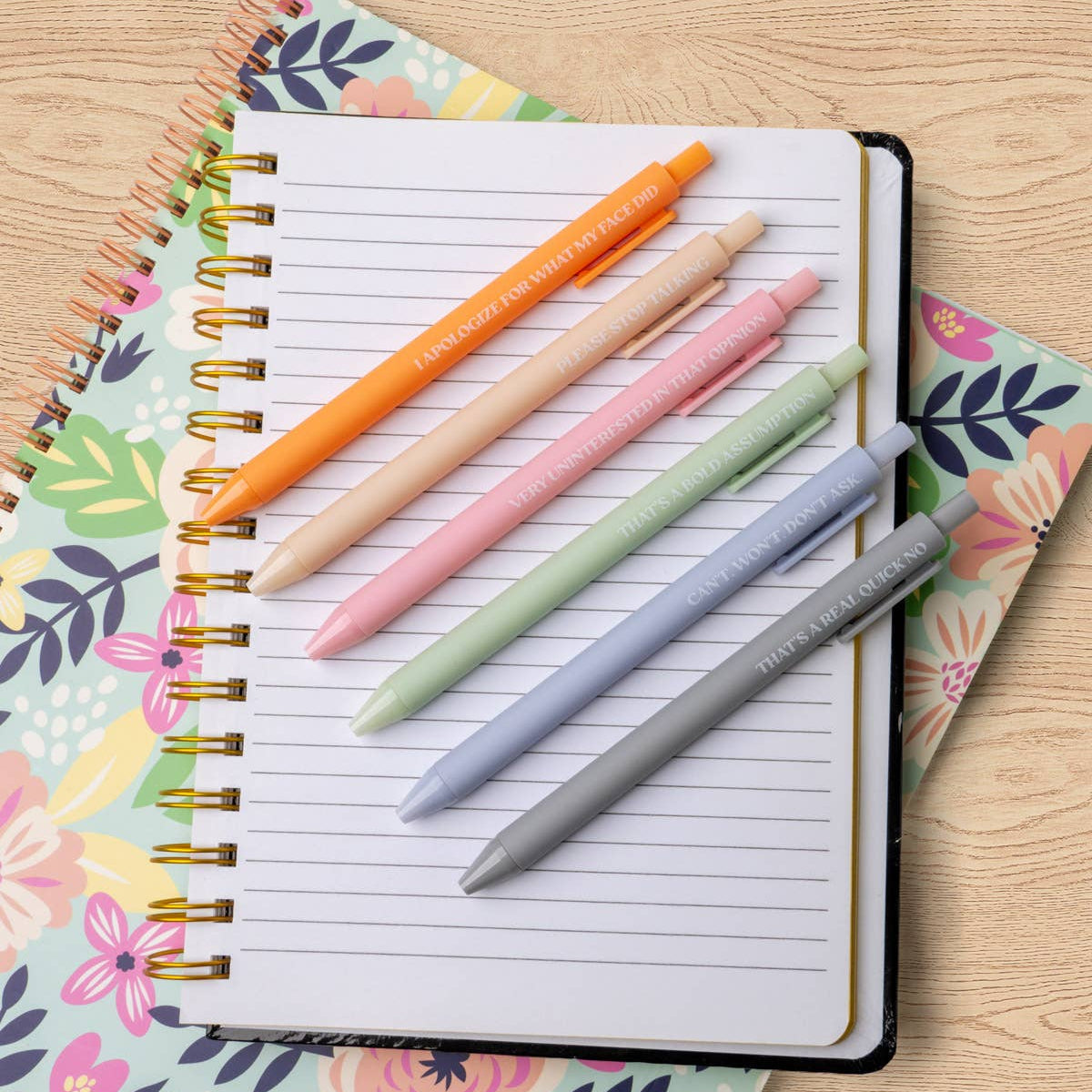 Mellow Monkey set of six pastel click ballpoint pens with funny quotes resting on a floral spiral notebook
