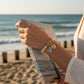 Mellow Monkey pearl bracelet featuring a hand-painted blue and white fish charm and gold shell on a beach background