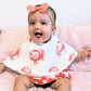Mellow Monkey baby wearing a white bamboo and cotton muslin bib with a pink crab print, sitting on a pink surface.