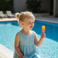A toddler in a blue dress holding a Mellow Monkey popsicle-shaped glittery squeeze toy by a swimming pool.