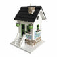 Mellow Monkey Primrose Cottage birdhouse with white exterior, black roof, green trim, and floral motif. Features river rock accents and hanging planters.