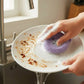 Mellow Monkey Good News Bears scrubby sponge in soft purple, with cute bear face, cleaning a dirty plate.