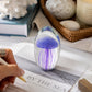 Mellow Monkey glass jellyfish paperweight in purple on a desk with a hand writing on a coastal themed notebook