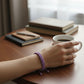 Mellow Monkey purple woven cotton nautical bracelet on a wrist holding a coffee mug at a desk.