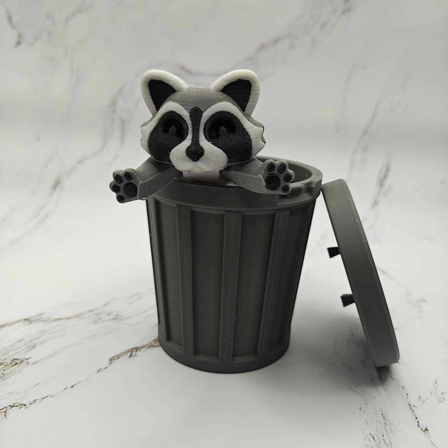 Mellow Monkey articulated raccoon fidget toy emerging from a grey 3D printed plastic trash can on a marble surface.