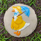 Mellow Monkey Rain Goose stepping stone featuring a white goose in a yellow raincoat and blue umbrella on stone base