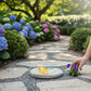 Mellow Monkey Rain Goose stepping stone plaque with yellow raincoat design placed on a garden path near purple flowers
