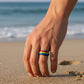 Mellow Monkey rainbow striped stainless steel ring worn on a finger against a sandy beach background.