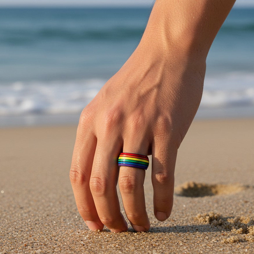 Mellow Monkey rainbow striped stainless steel ring worn on a finger against a sandy beach background.