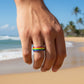 Mellow Monkey rainbow striped stainless steel ring worn on a finger against a sunny beach background.