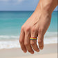 Mellow Monkey rainbow striped stainless steel ring worn on a finger against a blurred beach background.