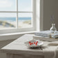 Mellow Monkey red enamel lobster earrings resting in a white shell dish on a wooden table by a beach view window.