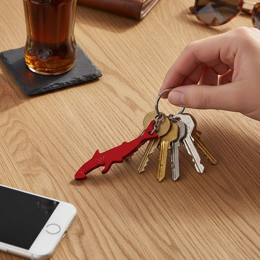 Mellow Monkey red aluminum shark-shaped bottle opener keychain held by a hand with keys on a wooden table.
