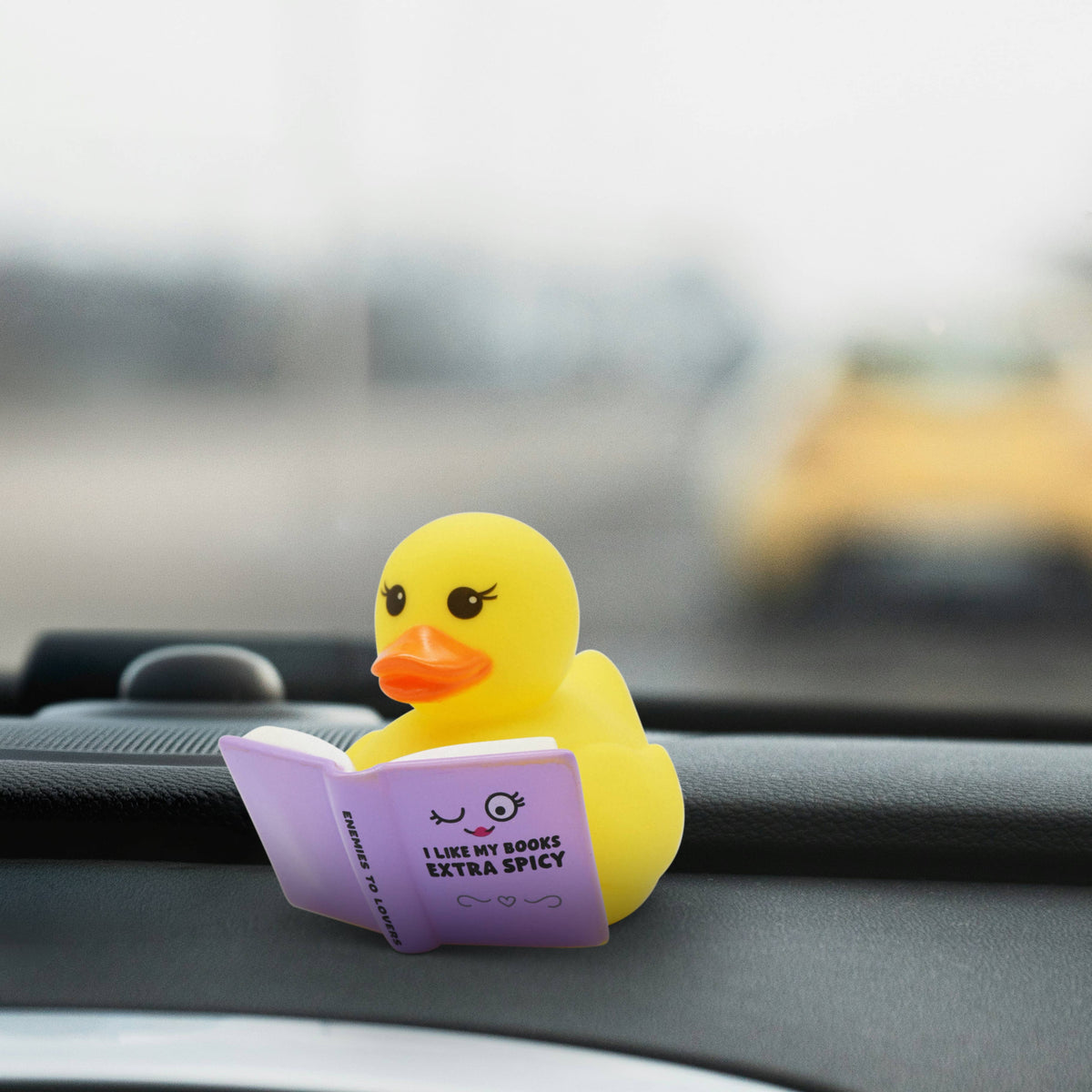 Mellow Monkey yellow rubber duck reading a purple book titled I Like My Books Extra Spicy on a car dashboard