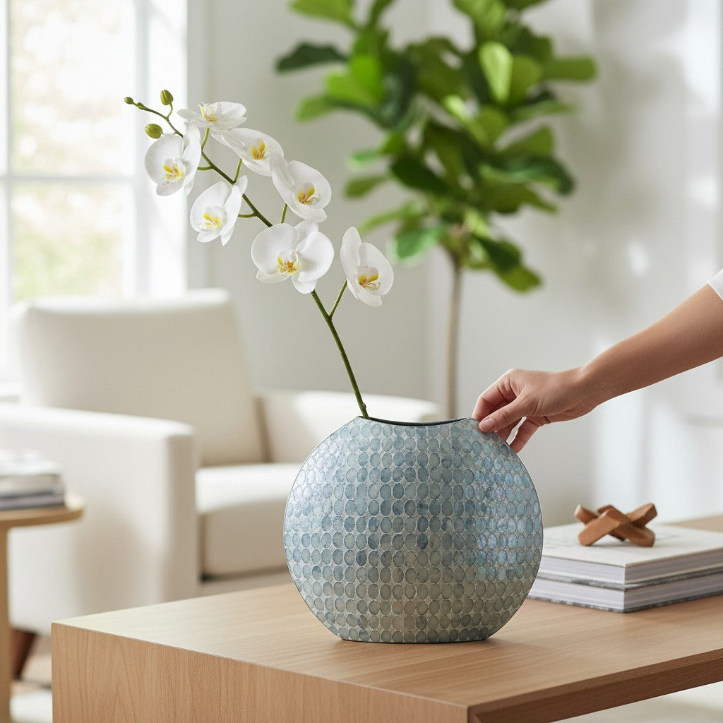 Mellow Monkey round blue capiz shell vase with iridescent texture holding white orchid stems on a wooden table.