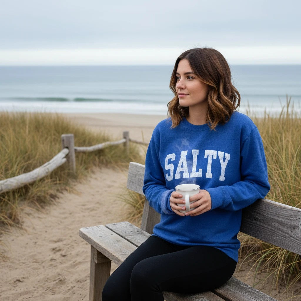 Mellow Monkey Salty Graphic Sweatshirt in heather royal blue, featuring a crewneck and white "SALTY" graphic. Model sitting on a bench by the beach.