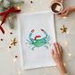 Mellow Monkey white cotton flour sack towel featuring a watercolor blue crab wearing a Santa hat on a festive table.