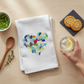 Mellow Monkey white cotton flour sack towel featuring a colorful sea glass heart design on a wooden kitchen table.