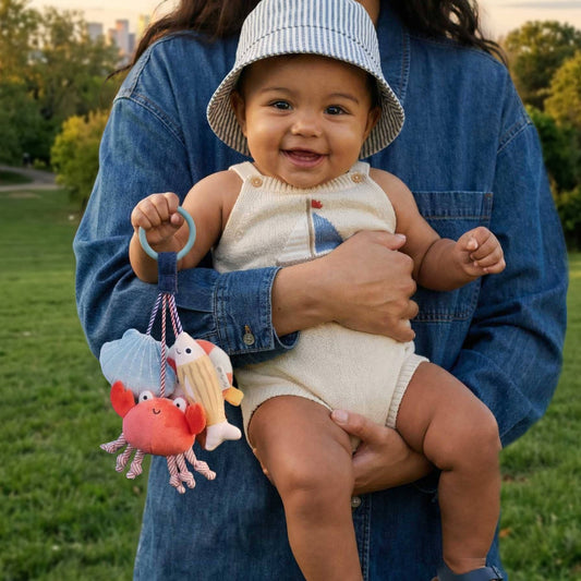 Mellow Monkey Itzy Travel Charms Seaside baby toy with crab, shell, and fish shapes held by a smiling baby outdoors.