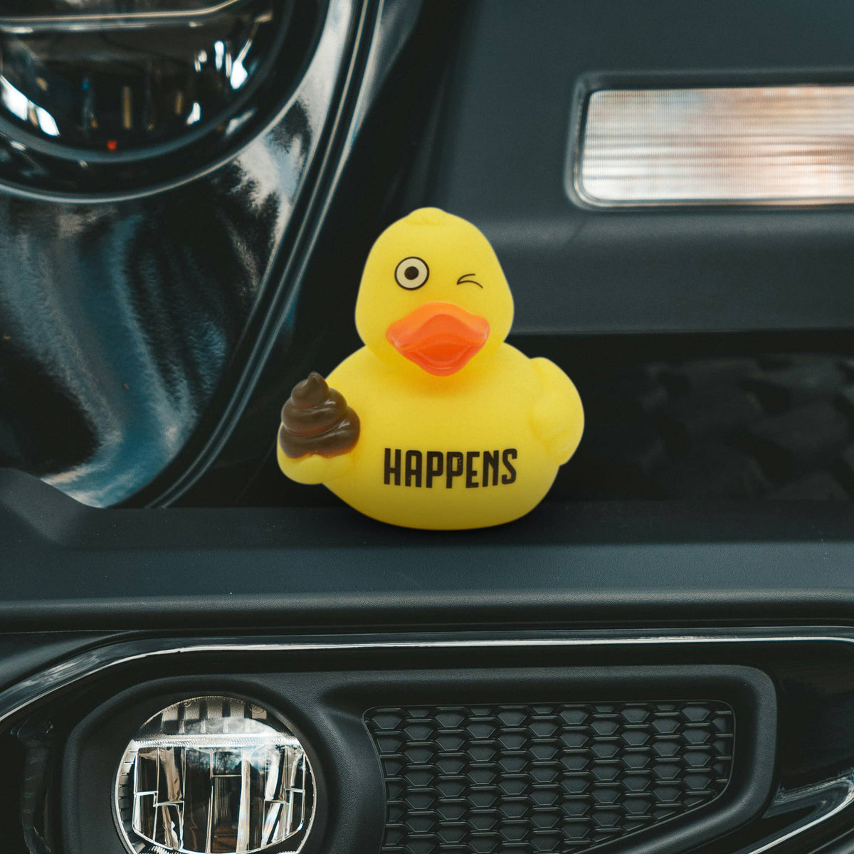 Mellow Monkey yellow rubber duck with a winking face, holding a brown poop emoji, sitting on a car bumper.