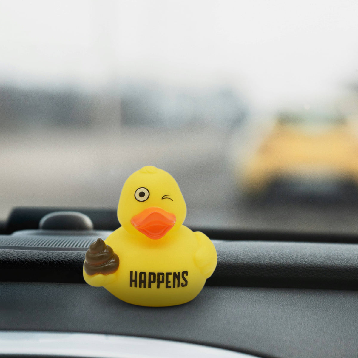 Mellow Monkey Shit Happens rubber duck with a winking expression, sitting on a car dashboard.