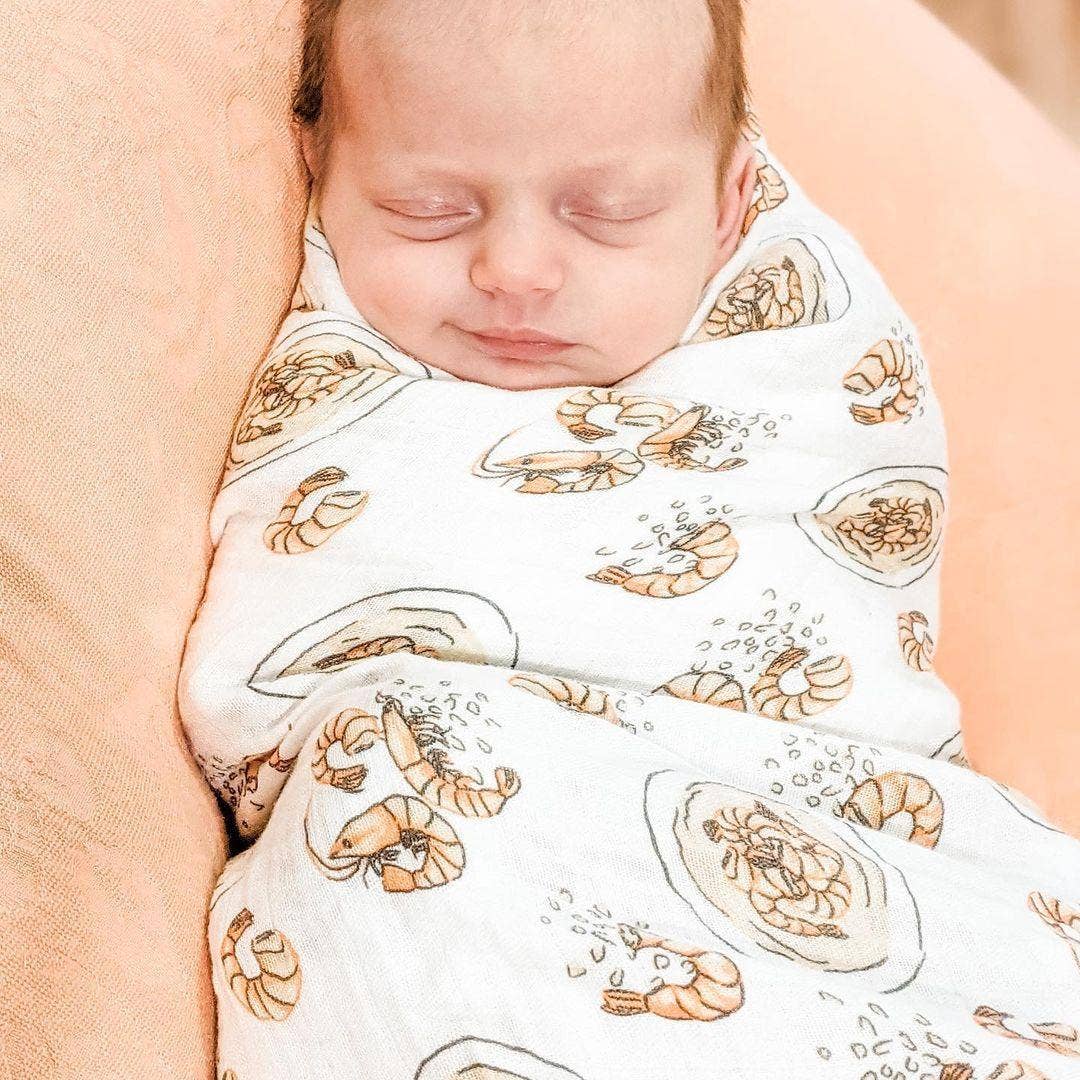 Mellow Monkey baby swaddled in a soft white muslin blanket featuring a shrimp and grits print