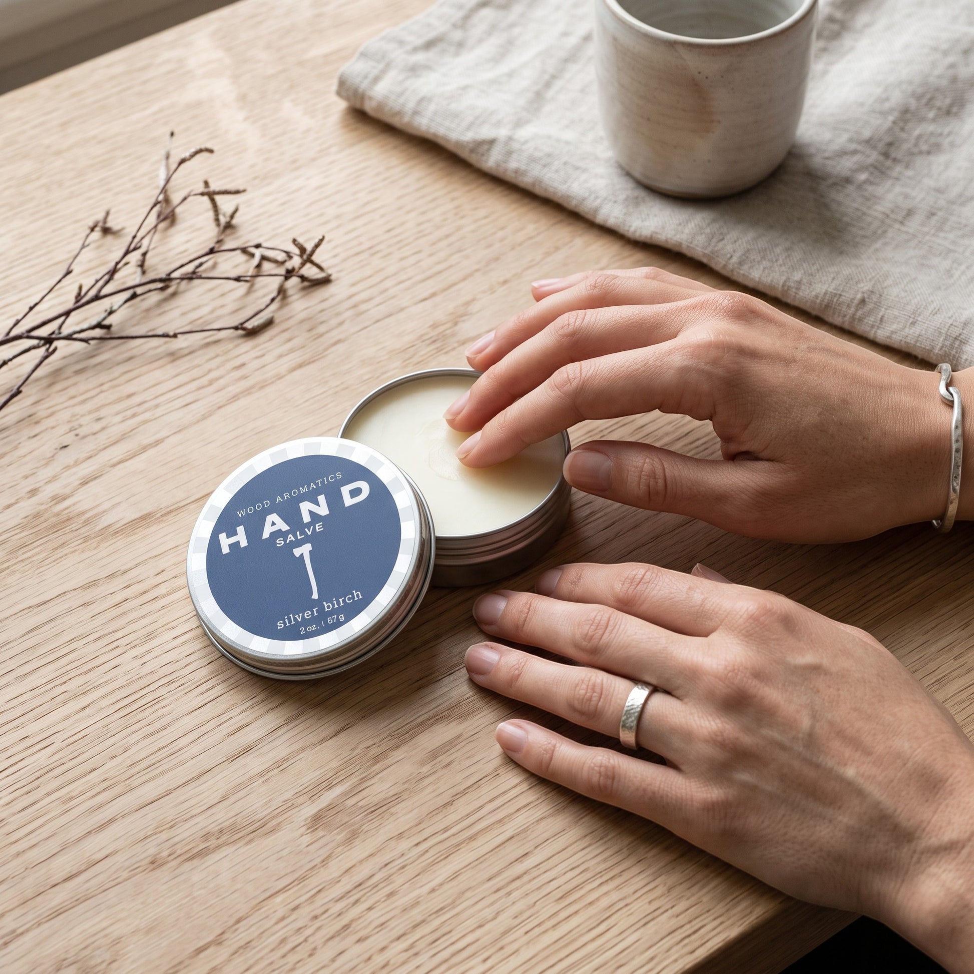 Mellow Monkey Wood Aromatics Silver Birch hand salve in a metal tin being applied by hand on a wooden table.