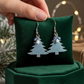 Mellow Monkey silver glittery Christmas tree earrings with surgical steel wires displayed on a green velvet cushion.