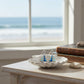 Mellow Monkey silver sailboat earrings with blue enamel, displayed in a white shell dish by a coastal window.