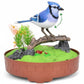 Mellow Monkey motion-activated blue and white singing bird toy perched on a branch with faux greenery in a brown base.